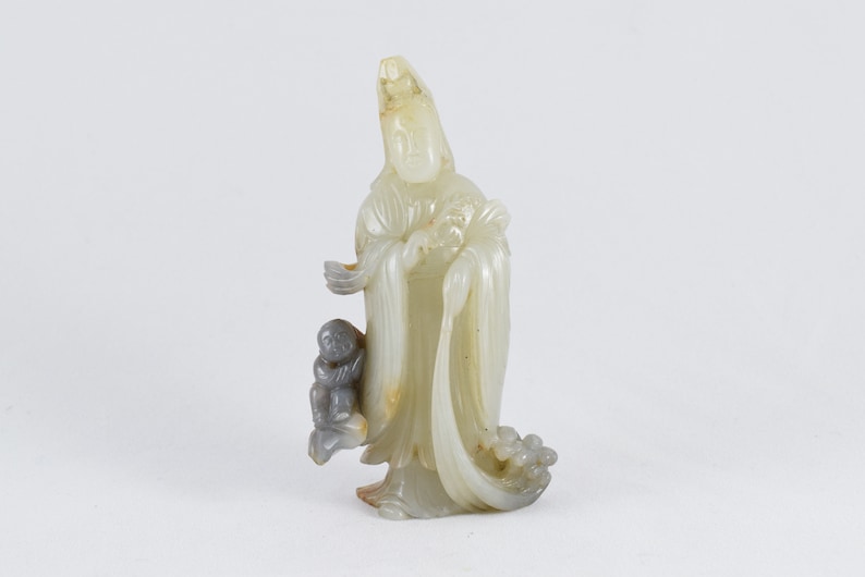 Puede incluir: Una escultura de jade blanco de una figura femenina con un ni&ntilde;o a su lado. La figura lleva una t&uacute;nica larga y una corona en la cabeza. La escultura est&aacute; tallada con un estilo detallado e intrincado.