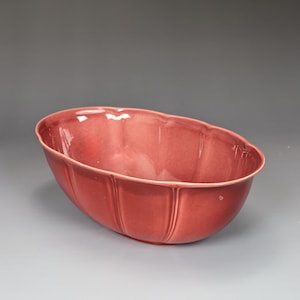 May include: A glossy, oval-shaped, red ceramic bowl. The bowl has a scalloped rim and vertical ribbed detailing on the sides. The interior of the bowl is smooth and reflects light. The bowl is likely for decorative or serving purposes.