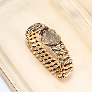 May include: A gold-toned expandable bracelet featuring a series of interlocking, textured metal links and a central heart-shaped clasp. The heart clasp is decorated with an engraved floral pattern and a smooth, oval-shaped center panel.