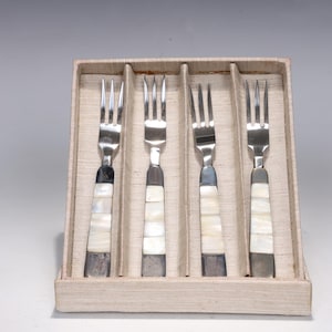 May include: A set of four silver-toned metal forks with handles inlaid with rectangular pieces of iridescent white mother-of-pearl. The forks are arranged vertically and held in place within a light beige, textured cardboard presentation box.