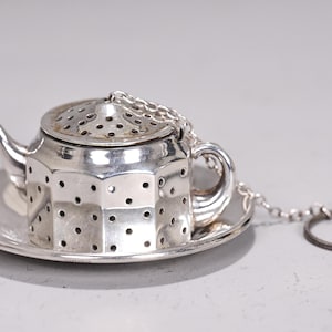 Könnte beinhalten: Silberner Tee-Ei in Teekannenform mit passender Untertasse. Der Aufgussbehälter hat einen perforierten Körper und Deckel, einen kleinen Ausguss und einen Griff. Eine Kette verbindet den Aufgussbehälter mit einem Ring. Zum Aufbrühen von losem Tee.