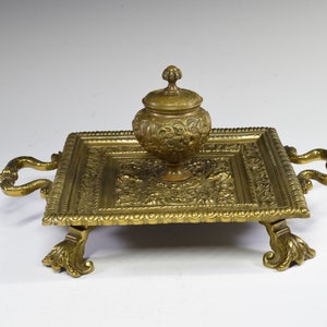 Può includere: Un calamaio in ottone dorato con un coperchio e dettagli decorativi. Il calamaio è posto su un vassoio quadrato con dettagli decorativi e quattro piedini. Il vassoio ha due maniglie sui lati.