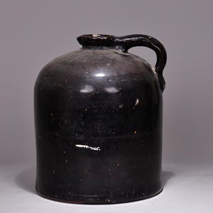 May include: A dark brown stoneware jug with a rounded body and a curved handle. The jug has a glossy finish and appears to be antique. The jug is likely used for storing liquids.