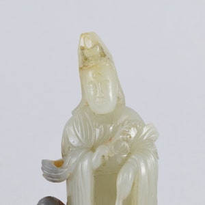 Puede incluir: Una escultura de jade blanco de una figura femenina que lleva una larga t&uacute;nica y una corona. La figura sostiene un abanico en su mano derecha y un rollo en su mano izquierda.