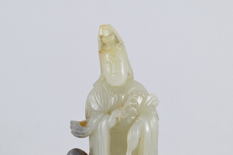 Puede incluir: Una escultura de jade blanco de una figura femenina que lleva una larga t&uacute;nica y una corona. La figura sostiene un abanico en su mano derecha y un rollo en su mano izquierda.