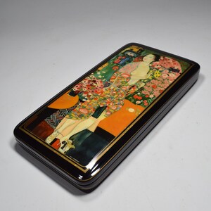 May include: A black lacquered wooden box with a colorful painting of a woman in a floral dress. The painting is in the style of Gustav Klimt.