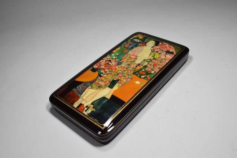 May include: A black lacquered wooden box with a colorful painting of a woman in a floral dress. The painting is in the style of Gustav Klimt.