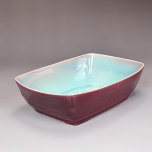 May include: A rectangular ceramic bowl with a burgundy exterior and a turquoise interior. The bowl has a slightly curved shape and a glossy finish. The exterior features horizontal ridges near the base.