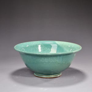 May include: A ceramic bowl featuring a glossy, variegated teal and turquoise glaze with a smooth, rounded rim. The bowl has a wide, open basin that tapers down to a circular footed base, revealing a thin, dark brown unglazed edge at the bottom.