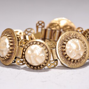 May include: A vintage gold-toned bracelet with large, round, cream-colored faux pearl centerpieces. The bracelet features ornate filigree detailing and a clasp. The bracelet is a statement piece of jewelry.