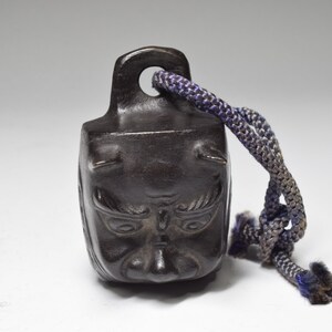 May include: A small, dark brown, bell-shaped object with a carved face and horns. The bell has a dark blue cord attached to it.