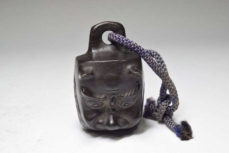 May include: A small, dark brown, bell-shaped object with a carved face and horns. The bell has a dark blue cord attached to it.