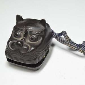 May include: A dark brown wooden hand-held bell shaped like a demon's face with a blue and gray rope attached.