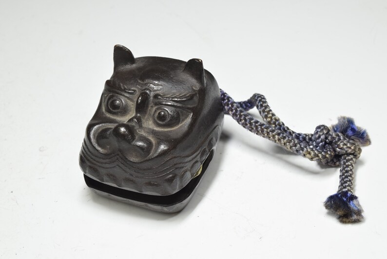 May include: A dark brown wooden hand-held bell shaped like a demon's face with a blue and gray rope attached.