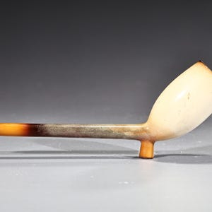Puede incluir: Una pipa de tabaco vintage con un cuenco color crema y un tallo largo y delgado. El tallo pasa de un marrón oscuro a un color ámbar más claro. La pipa tiene un diseño clásico y curvado.