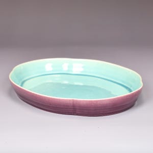 May include: An oval-shaped ceramic serving dish with a light teal interior and a deep purple exterior. The rim has a wavy edge. The dish is likely used for serving food or as a decorative piece.