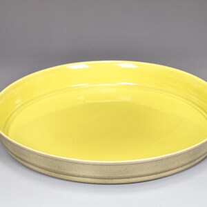 May include: A round, ceramic serving dish with a bright yellow interior and a light beige exterior. The dish has a wide, shallow design with a slightly raised rim. The interior is smooth and glossy, while the exterior has a textured appearance.