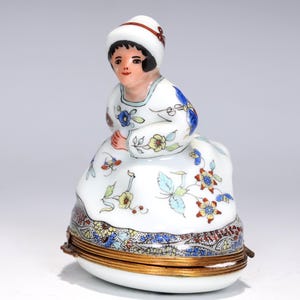 May include: A porcelain figurine of a seated woman in a white dress and hat, adorned with floral patterns in blue, yellow, and red. The base is a hinged box with a gold-colored rim and a decorative border.