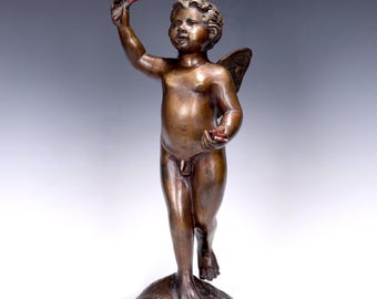 May include: Bronze statue of a cherub with wings, holding a red object in each hand. The cherub stands on a round base with a dark rim. The statue has a detailed finish and is set against a gradient background.