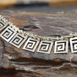 Könnte beinhalten: Silberne Kettenkette mit geometrischem Muster. Die Kette besteht aus kleinen, miteinander verbundenen Quadraten mit einem griechischen Schlüsselmuster.