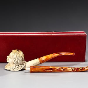 Puede incluir: Una pipa de tabaco tallada con una cara detallada en el cuenco, un tallo de color crema y una boquilla curva marrón y ámbar. La pipa descansa frente a una caja roja.