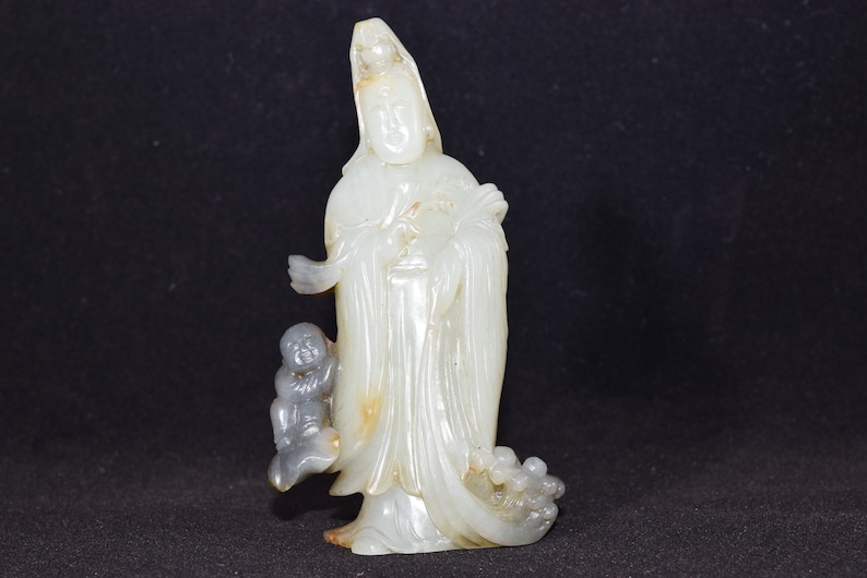 Puede incluir: Una escultura de jade blanco de una figura femenina de pie con un ni&ntilde;o a su lado. La figura lleva una t&uacute;nica larga y tiene una expresi&oacute;n serena. La escultura est&aacute; tallada y pulida de forma intrincada.