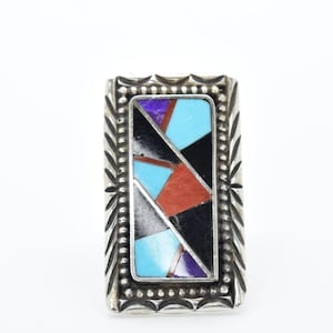 Puede incluir: Anillo rectangular de plata con incrustaciones de mosaico de formas geométricas turquesas, negras, rojas y moradas. El anillo tiene un borde decorativo con un patrón detallado.