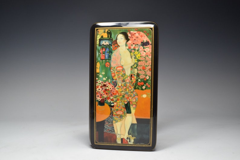 May include: A black lacquered wooden box with a colorful painting of a woman standing in a garden with flowers. The woman is wearing a long, patterned dress and has long dark hair. The box is rectangular and has a hinged lid.