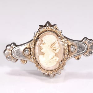 May include: A silver-toned metal brooch featuring an oval central cameo depicting a profile of a person. The frame is ornate with textured detailing and two small circular accents on either side of the central oval, finished with a polished sheen.