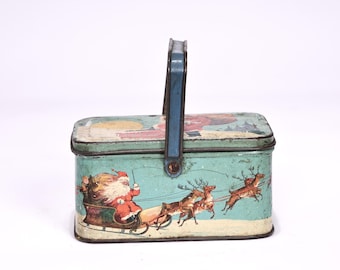 May include: Vintage rectangular tin box with a blue handle and a festive Christmas theme. The box features Santa Claus in his sleigh being pulled by reindeer against a light blue background. The box is likely used for storage or as a decorative item.
