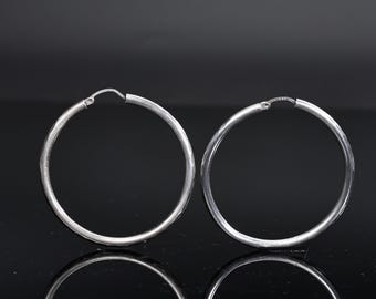 May include: A pair of silver hoop earrings. The earrings are circular with a simple, polished design. The hoops are displayed against a dark background, with their reflections visible below.