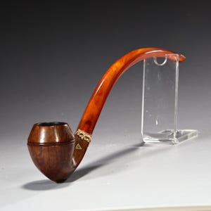 Puede incluir: Una pipa de tabaco curvada con un cuenco marrón oscuro y una boquilla curvada marrón claro. La boquilla tiene una banda dorada decorativa cerca del cuenco. La pipa se exhibe en un soporte transparente.
