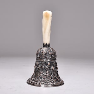 Puede incluir: Una campana de plata antigua con un mango de marfil. La campana presenta grabados en relieve detallados de figuras y rostros. El mango es de color crema claro. La campana es un objeto decorativo.