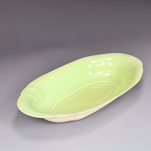 May include: A light green oval-shaped ceramic serving dish with a scalloped rim. The interior of the dish is a lighter shade of green, and the base is off-white. The dish has a glossy finish and is empty.