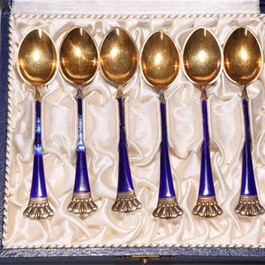 以下が含まれることがあります： ベルベット張りの箱に入った、青いエナメル製のハンドルが付いた銀製のティースプーン6本セット。スプーンは金メッキが施され、装飾的なデザインが施されています。