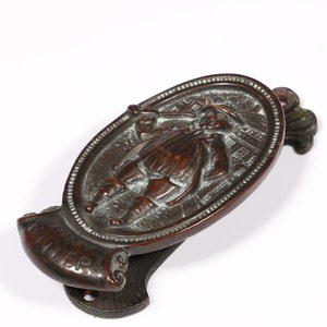 May include: A vintage bronze door knocker with an oval design featuring a raised image of a person in period clothing. The knocker has a scroll-shaped base with the word "Welcome" inscribed.