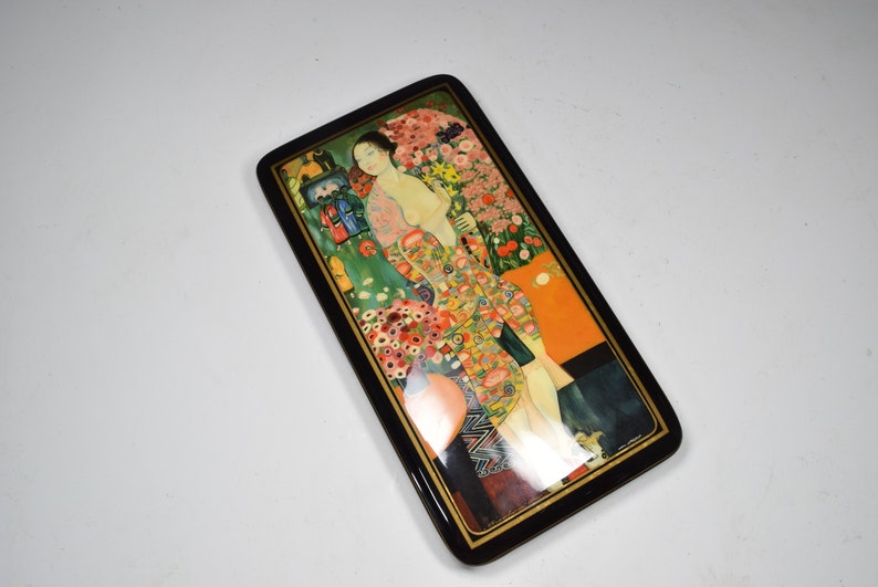 May include: A rectangular wooden box with a black lacquered finish and a colorful painted scene of a woman in a floral dress standing in a garden. The box has a gold trim around the edges.