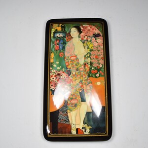 May include: A wooden box with a hinged lid featuring a colorful painting of a woman standing in a garden with flowers. The woman is wearing a patterned dress and has long dark hair.