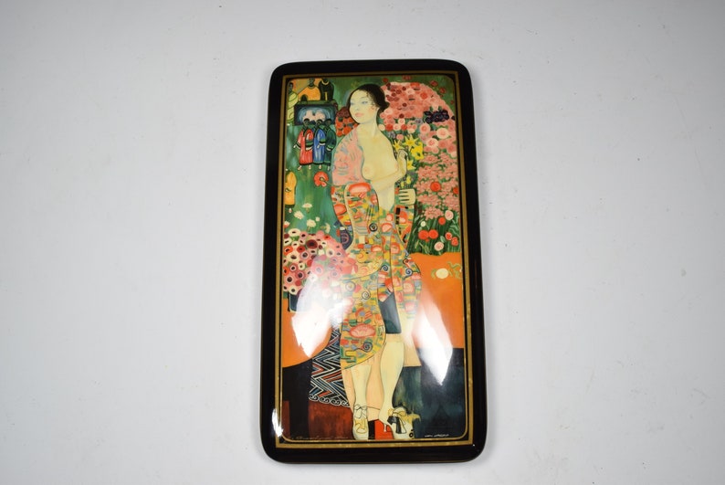May include: A wooden box with a hinged lid featuring a colorful painting of a woman standing in a garden with flowers. The woman is wearing a patterned dress and has long dark hair.