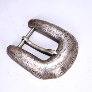 Könnte beinhalten: Silberfarbene Gürtelschnalle mit eingraviertem floralem Design. Die Schnalle hat eine gebogene Form mit einem Mittelsteg für den Gürtel. Das Metall hat ein leicht gealtertes Aussehen.