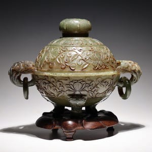 May include: A green jade incense burner with intricate floral carvings and two dragon-shaped handles. The burner sits on a wooden stand with three legs.