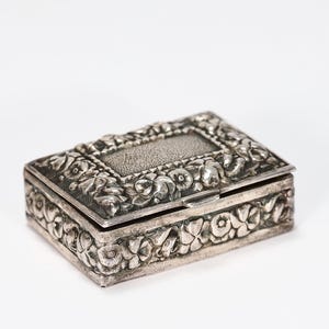 Puede incluir: Una caja rectangular de metal plateado con un intrincado patrón floral en relieve que cubre los lados y la tapa. La tapa incluye un panel central rectangular con un acabado de superficie texturizado y punteado, y un cierre de bisagra.