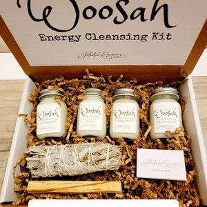 May include: A white box with the words "Woosah Energy Cleansing Kit" on the lid. The box contains four small candles, a bundle of sage, a wooden stick, and a small box of matches. The candles are labeled "Woosah Energy Cleansing" with different scents. The box is filled with brown paper shreds.
