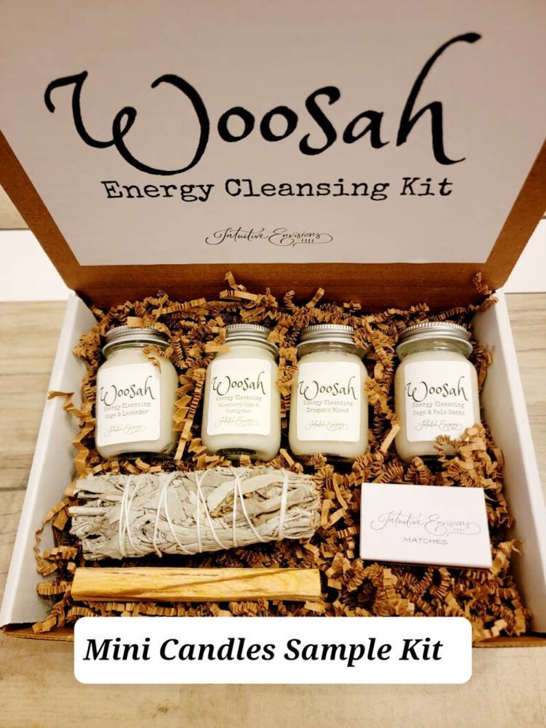 May include: A white box with the words "Woosah Energy Cleansing Kit" on the lid. The box contains four small candles, a bundle of sage, a wooden stick, and a small box of matches. The candles are labeled "Woosah Energy Cleansing" with different scents. The box is filled with brown paper shreds.