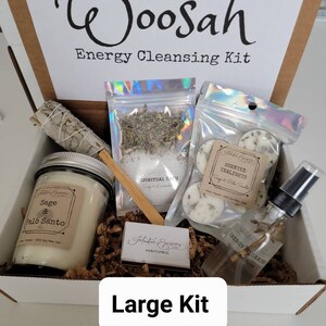 May include: A white box with the words "Woosah Energy Cleansing Kit" printed on the front. The box contains a variety of items, including a sage and palo santo candle, a bag of spiritual bath salts, a bag of scented tealights, a bottle of energy clearing spray, and a box of matches. The box is labeled "Large Kit".