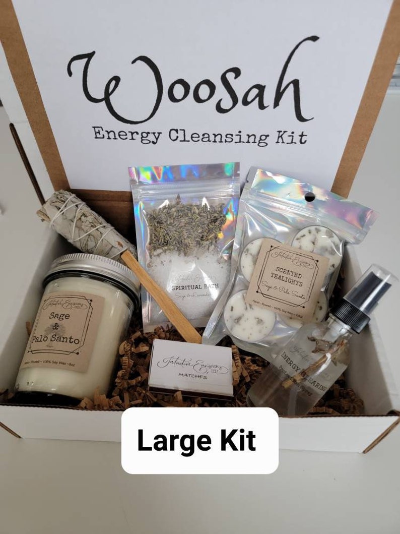 May include: A white box with the words "Woosah Energy Cleansing Kit" printed on the front. The box contains a variety of items, including a sage and palo santo candle, a bag of spiritual bath salts, a bag of scented tealights, a bottle of energy clearing spray, and a box of matches. The box is labeled "Large Kit".