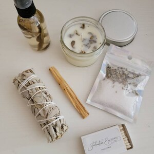 May include: A white table with a variety of items for a spiritual cleansing ritual. The items include a clear glass bottle with a spray top, a white candle in a glass jar, a bundle of white sage, a wooden stick, a small bag of dried lavender, and a box of matches labeled "Intuitive Expressions".