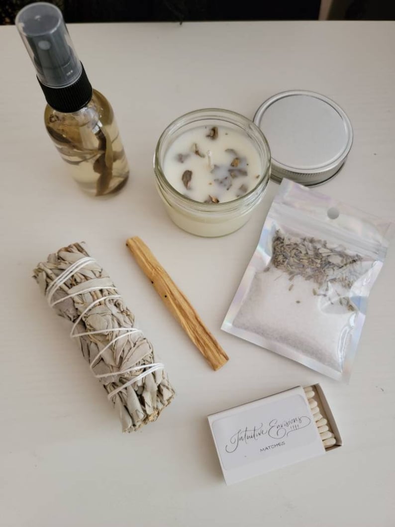 May include: A white table with a variety of items for a spiritual cleansing ritual. The items include a clear glass bottle with a spray top, a white candle in a glass jar, a bundle of white sage, a wooden stick, a small bag of dried lavender, and a box of matches labeled "Intuitive Expressions".