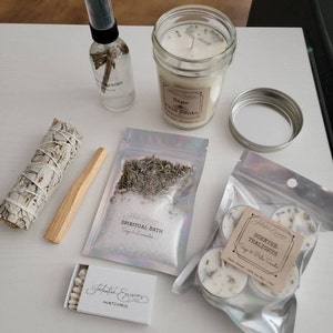 May include: A white sage smudge stick, palo santo stick, a small box of matches, a clear glass bottle with a spray nozzle, a jar candle with a white candle and a label that says "Sage & Palo Santo", a small bag of white bath salts with a label that says "Spiritual Bath Sage & Lavender", and a small bag of tea lights with a label that says "Scented Tealights Sage & Palo Santo".