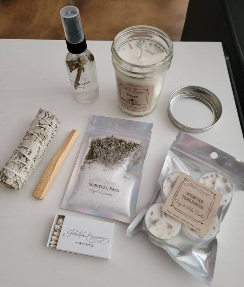 May include: A white sage smudge stick, palo santo stick, a small box of matches, a clear glass bottle with a spray nozzle, a jar candle with a white candle and a label that says "Sage & Palo Santo", a small bag of white bath salts with a label that says "Spiritual Bath Sage & Lavender", and a small bag of tea lights with a label that says "Scented Tealights Sage & Palo Santo".